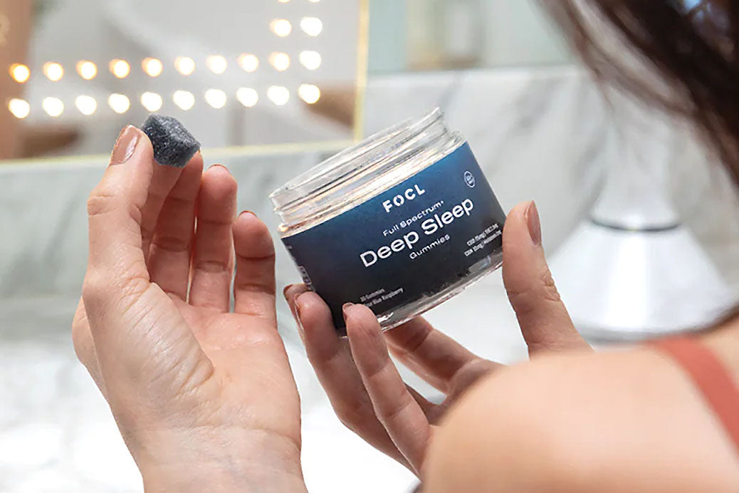 A woman taking a blue CBD gummy from a jar of FOCL Deep Sleep in front of a mirror.