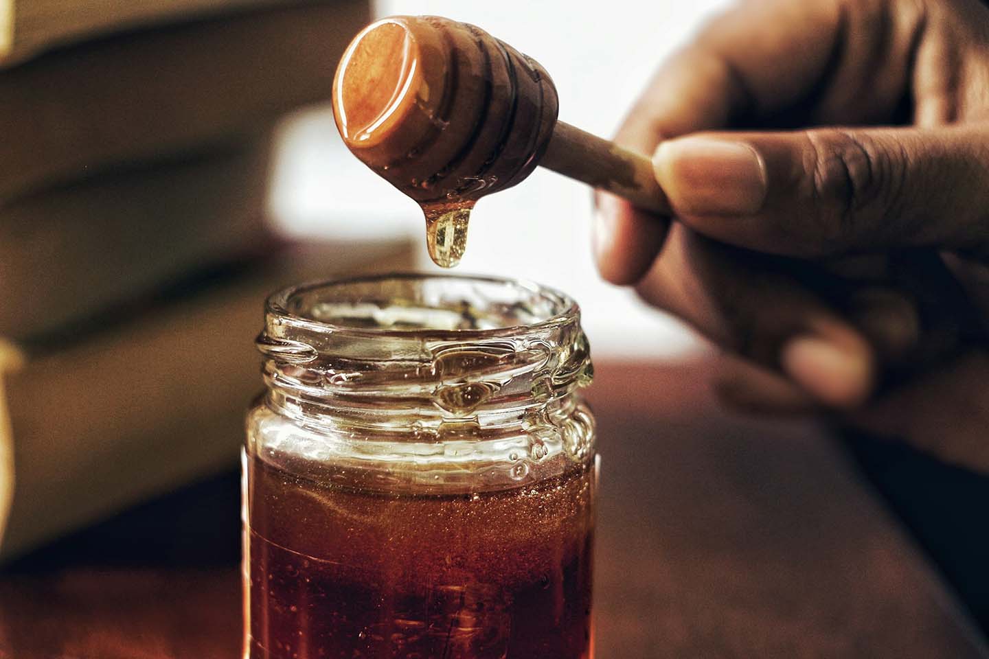 a hand holding up a honey dipper over a jar filled with amber syrup