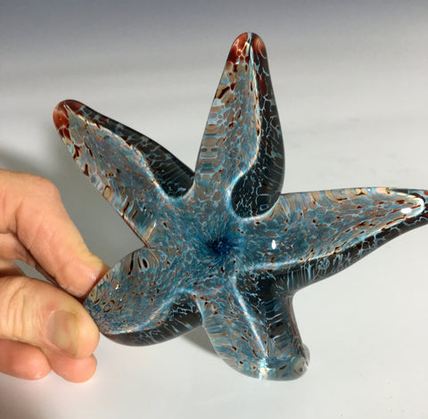 hand blown glass sea star or starfish with blue and copper red colors being held up with thumb and finger