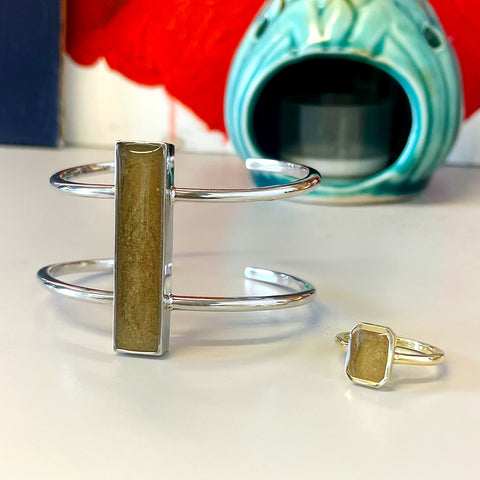 Close By Me's Bar Cremation Bracelet in Sterling Silver and Cushion Art Deco Cremation Ring in 14K Yellow Gold, both set with ashes from aquamation, resting on a grey surface.