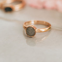 A close-up photo of Close By Me's Fleur De Lis Cremation Ring in 14K Rose Gold setting on a glass-like surface.