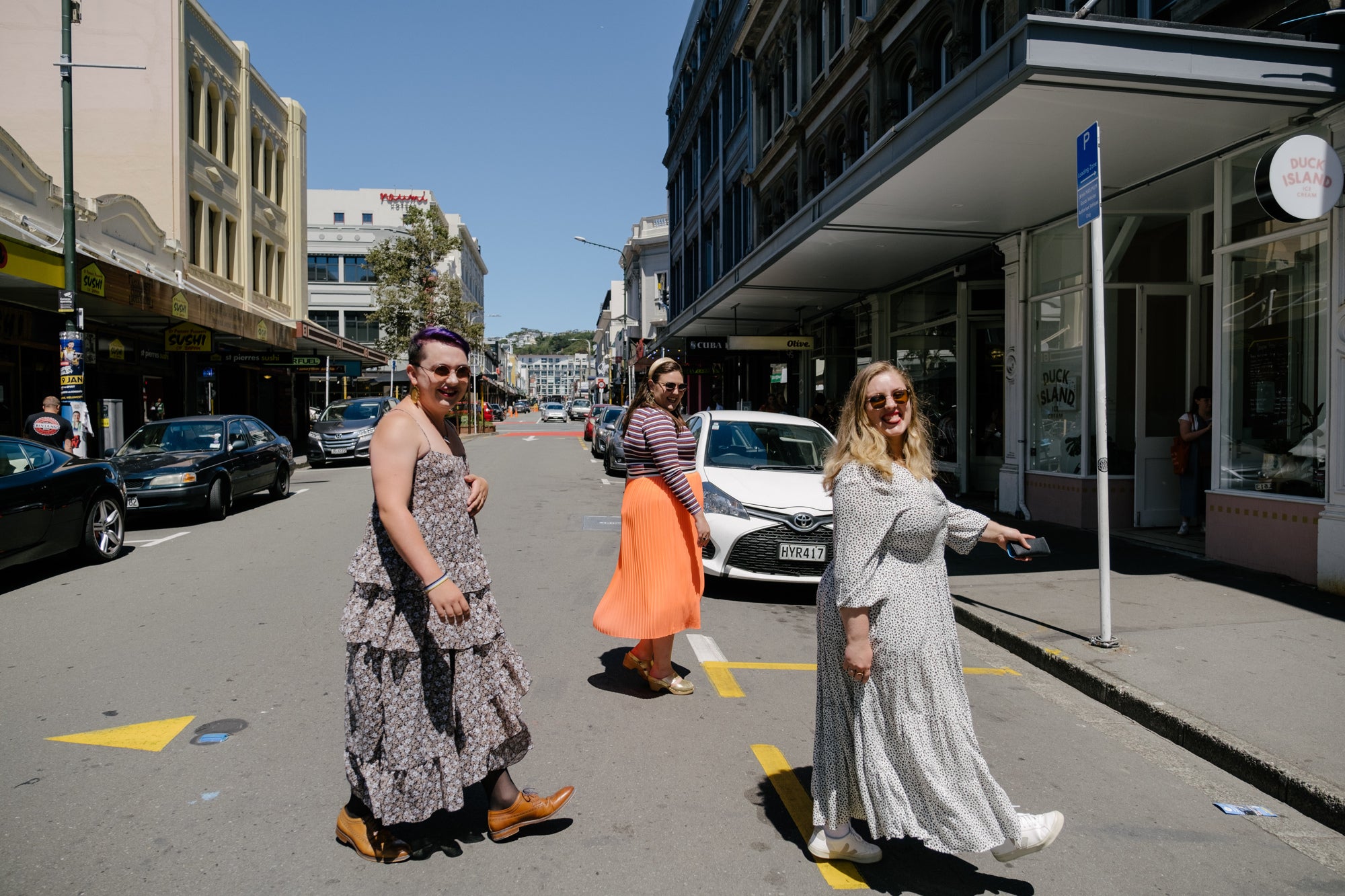 Maxwell, Lucy and Sasha all walking across Cuba Street. Maxwell is wearing a floral, floaty maxi, Sasha is wearing a purple stripey top, and Lucy is wearing a floor length white dress with small black leopard print spots on it. 