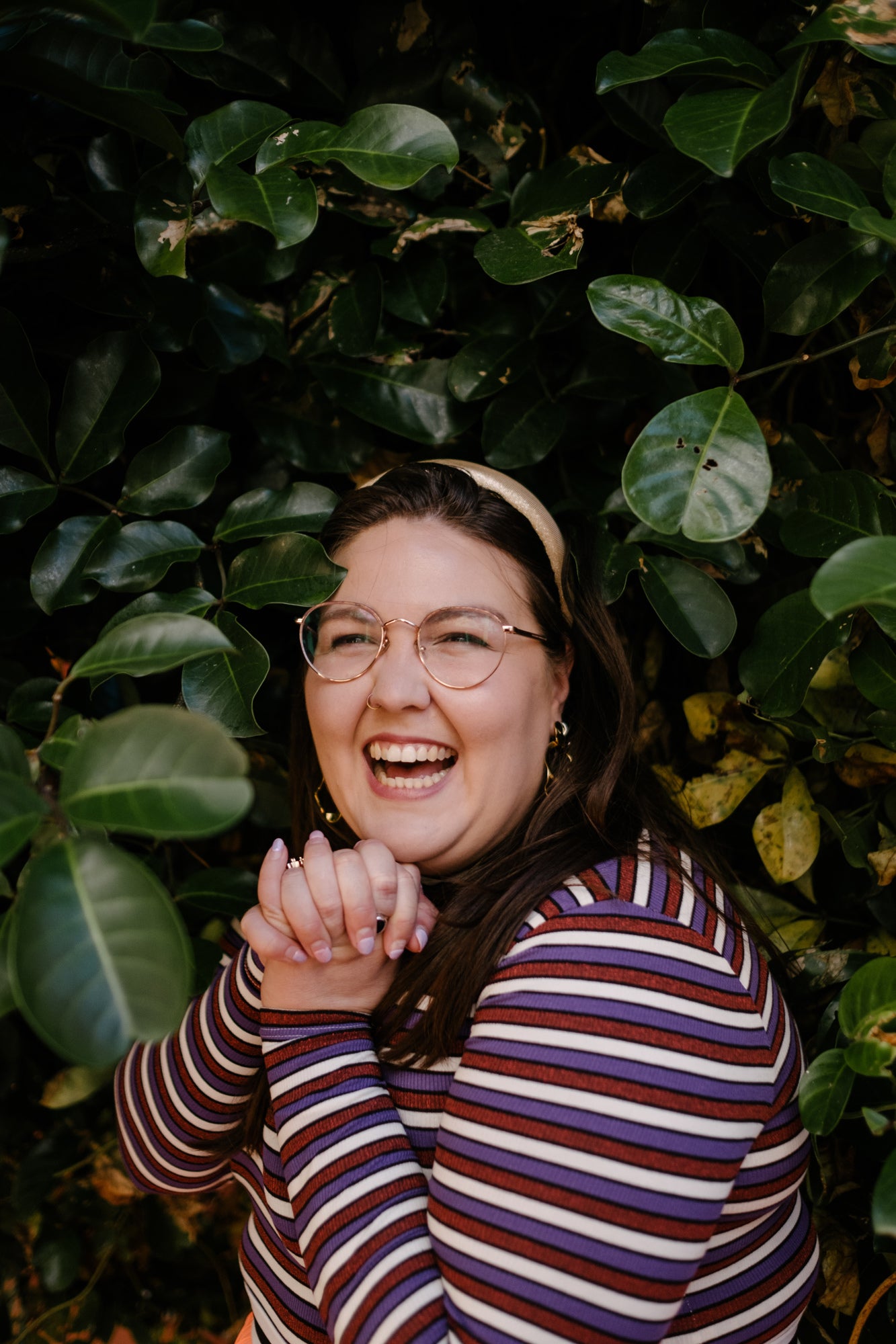 Sasha laughing in the midst of green foliage in a purple stripey top and glasses. 