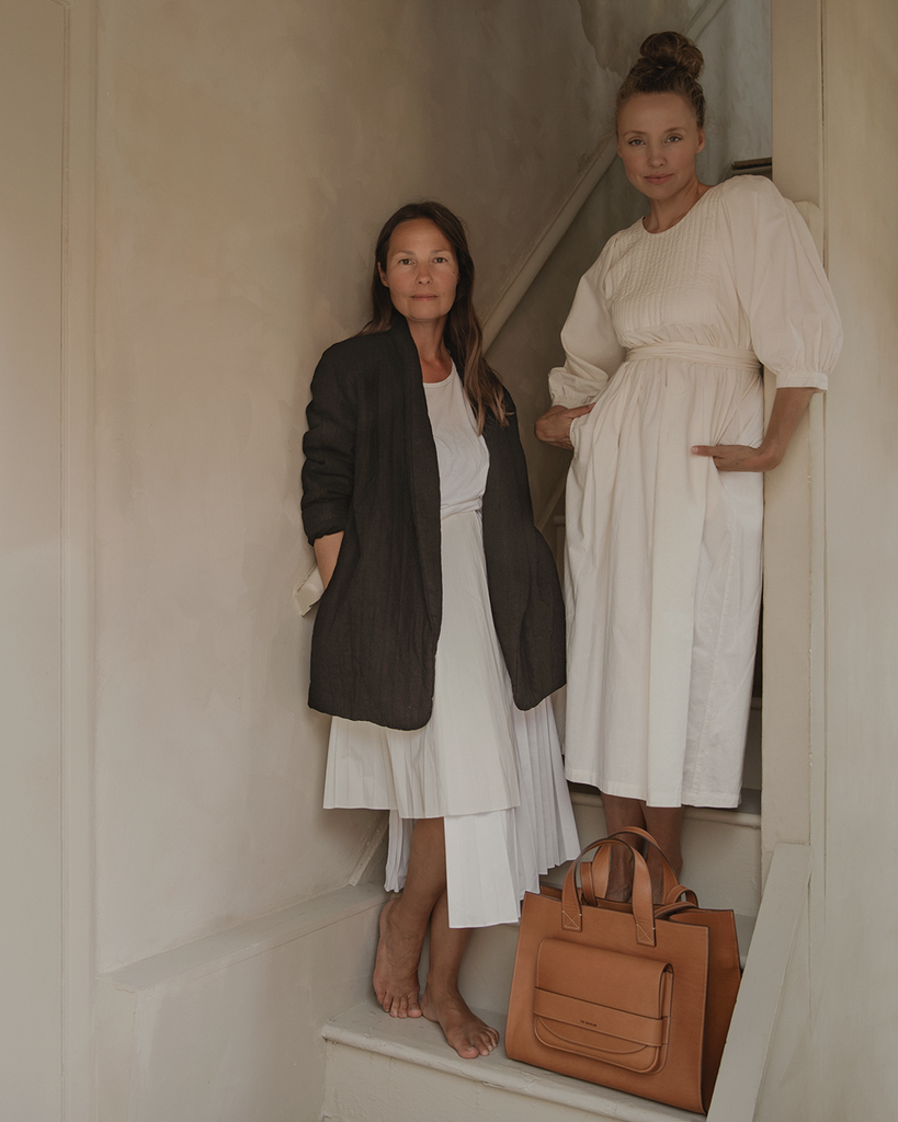 Hannah Lemholt and Carissa Wyten wearing quilted appareld from The Regular, standing on a staircase with a tan leather vegetable tanned leather 'Pioneer' tote bag