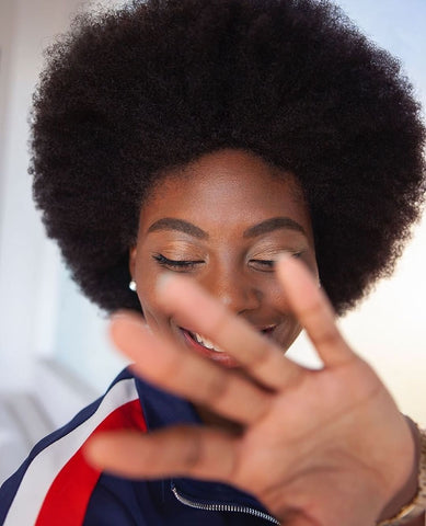 A Black girl with an afro smiling with her hand in front of her face