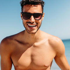 A shirtless young man wearing sunglasses smiling and standing at the beach with the ocean in the background.