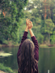 Woman with long brown hair is in nature near a lake and raises her hands in the air