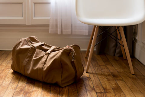 One brown leather duffle bag sitting on the wood floor next to a white stool, representing packing light