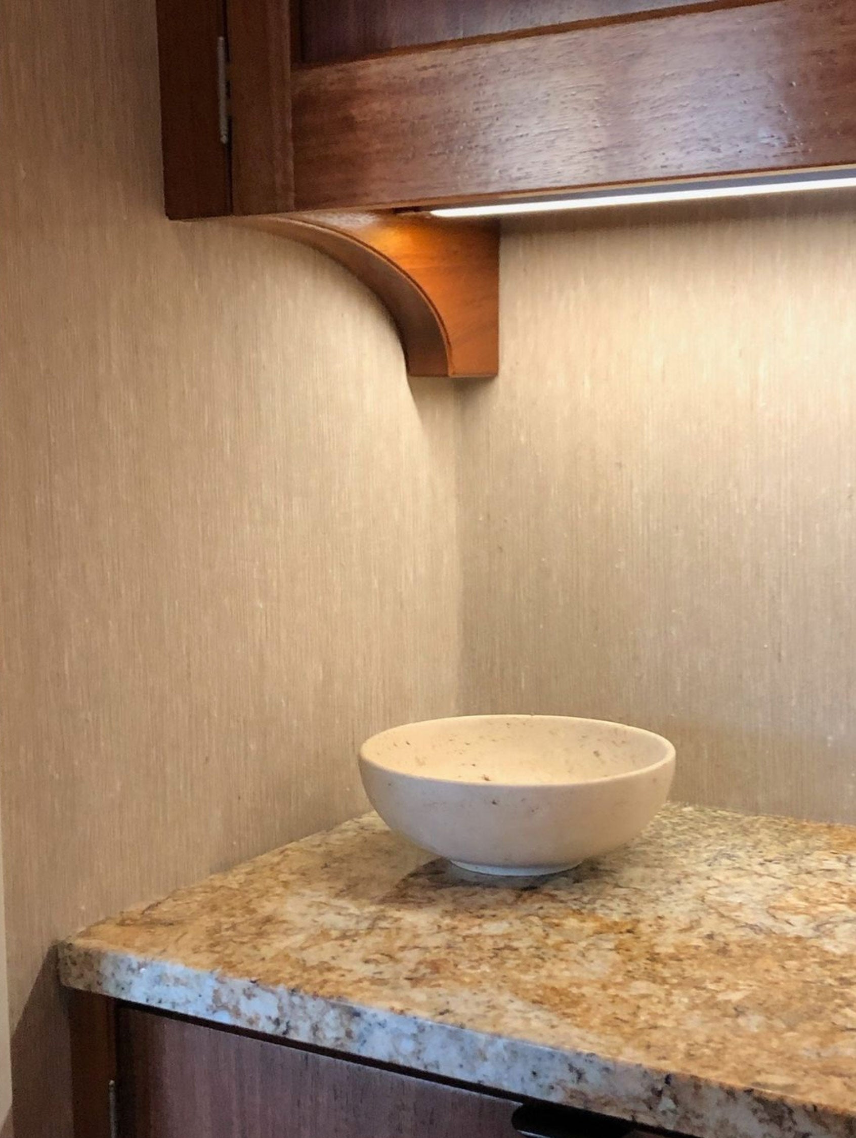 Closeup corner image of a neutral-toned natural wallcovering under a cabinet shelf. Small white bowl on countertop