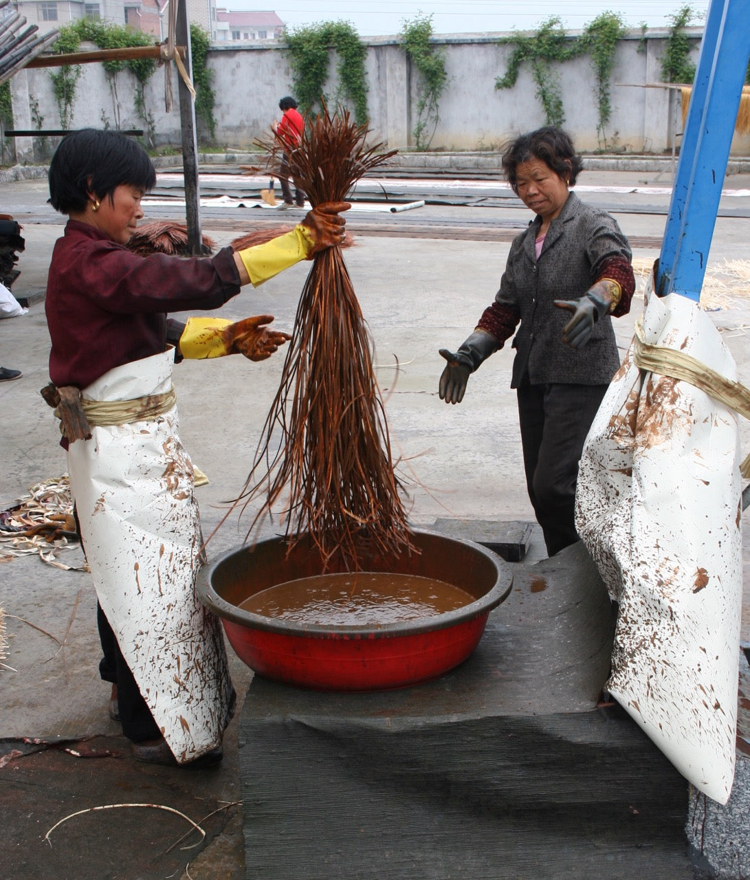 Two people hand-dipping long grass fibers in a pail of earth-red dye for production of earth-friendly natural wallcoverings