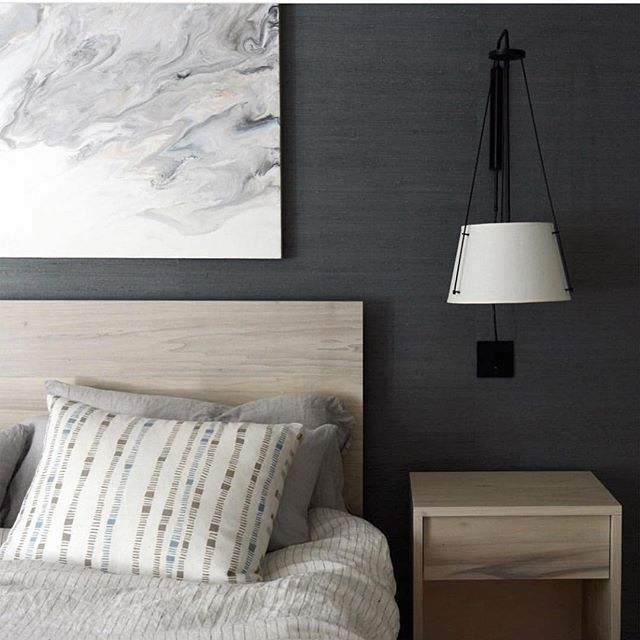 Closeup image of bed with light wooden headboard. A large white painting hangs behind on a dark charcoal colored natural grasscloth wallcovering