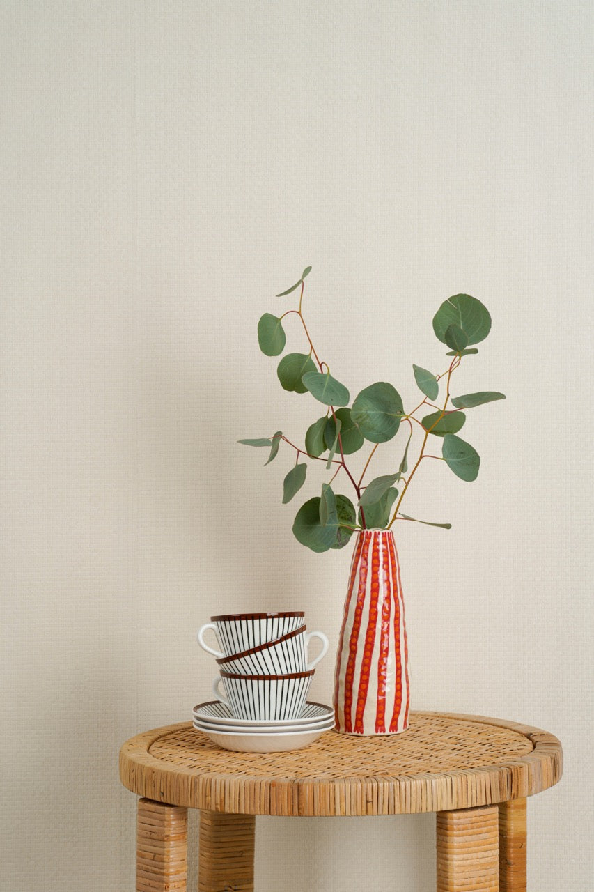 Red and white striped vase with eucalyptus leaves on a table next to a stack of teacups in front of white paperweave wallpaper