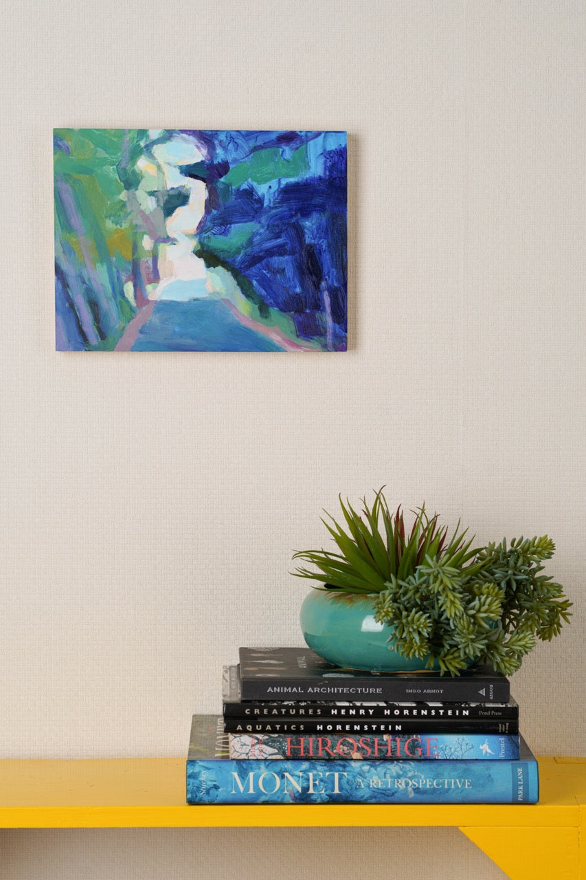 A colorful abstract painting hangs on a grasscloth wall above a stack of art books with a potted plant on top, creating a spring decor vignette.