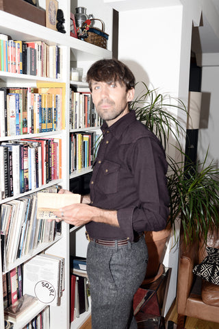 Dylan in front of a bookshelf, holding a book, wearing a burgundy western shirt, and gray tweed pants.