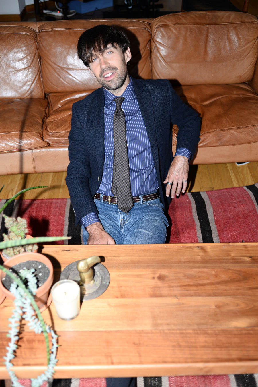 Dylan shot wearing jeans, a blue and white striped dress shirt, and a navy blazer sitting on the floor in front of a leather couch.