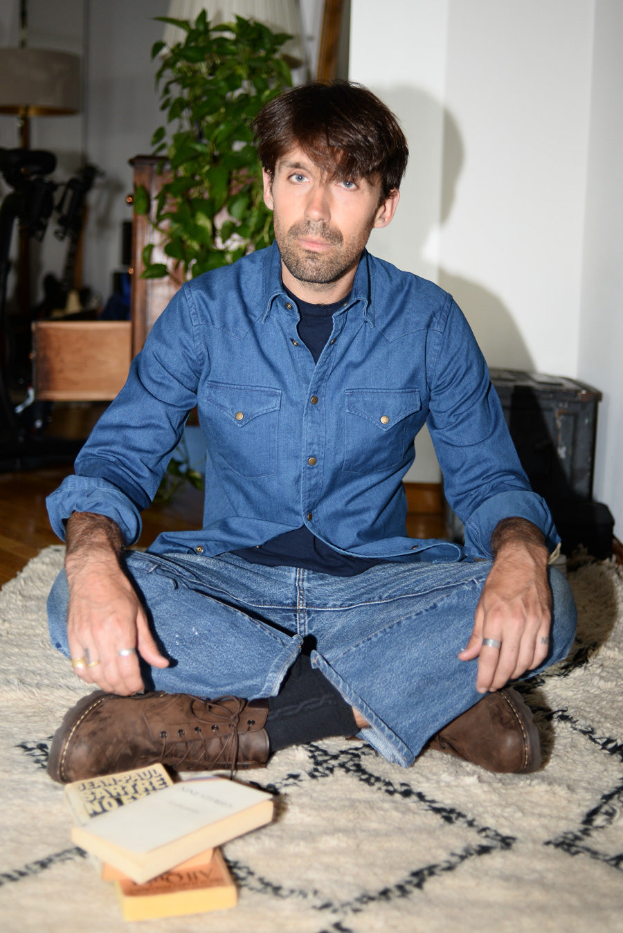 Dylan sits crossed-legged wearing jeans, a navy t-shirt, and a chambray western shirt.