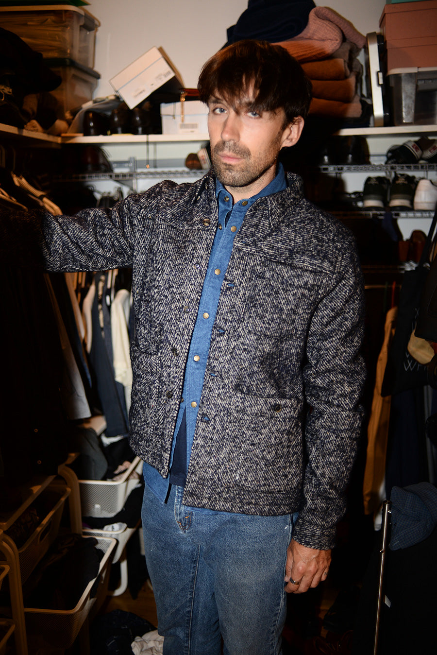 Dylan stands in a closet. He is wearing jeans, a chambray western shirt with a wool twill overshirt in navy.
