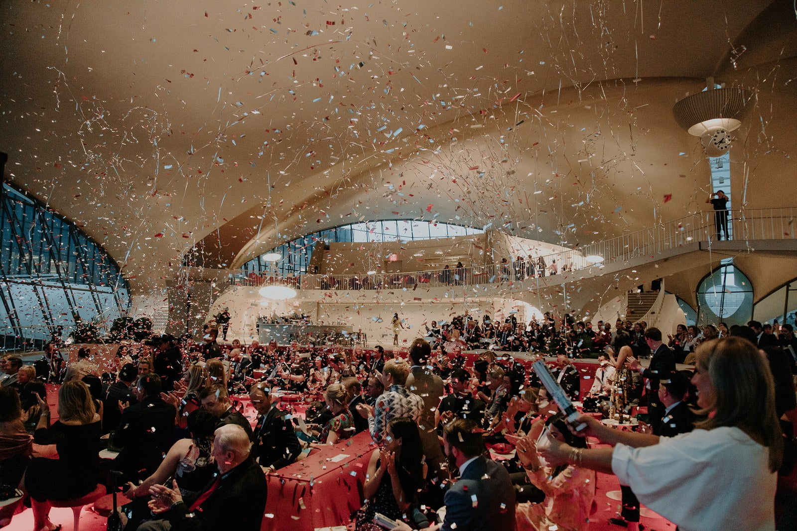 Wide shot of dozens of people celebrating at Perry and Adri's wedding at the TWA Hotel in NYC. There are streamers in the air.