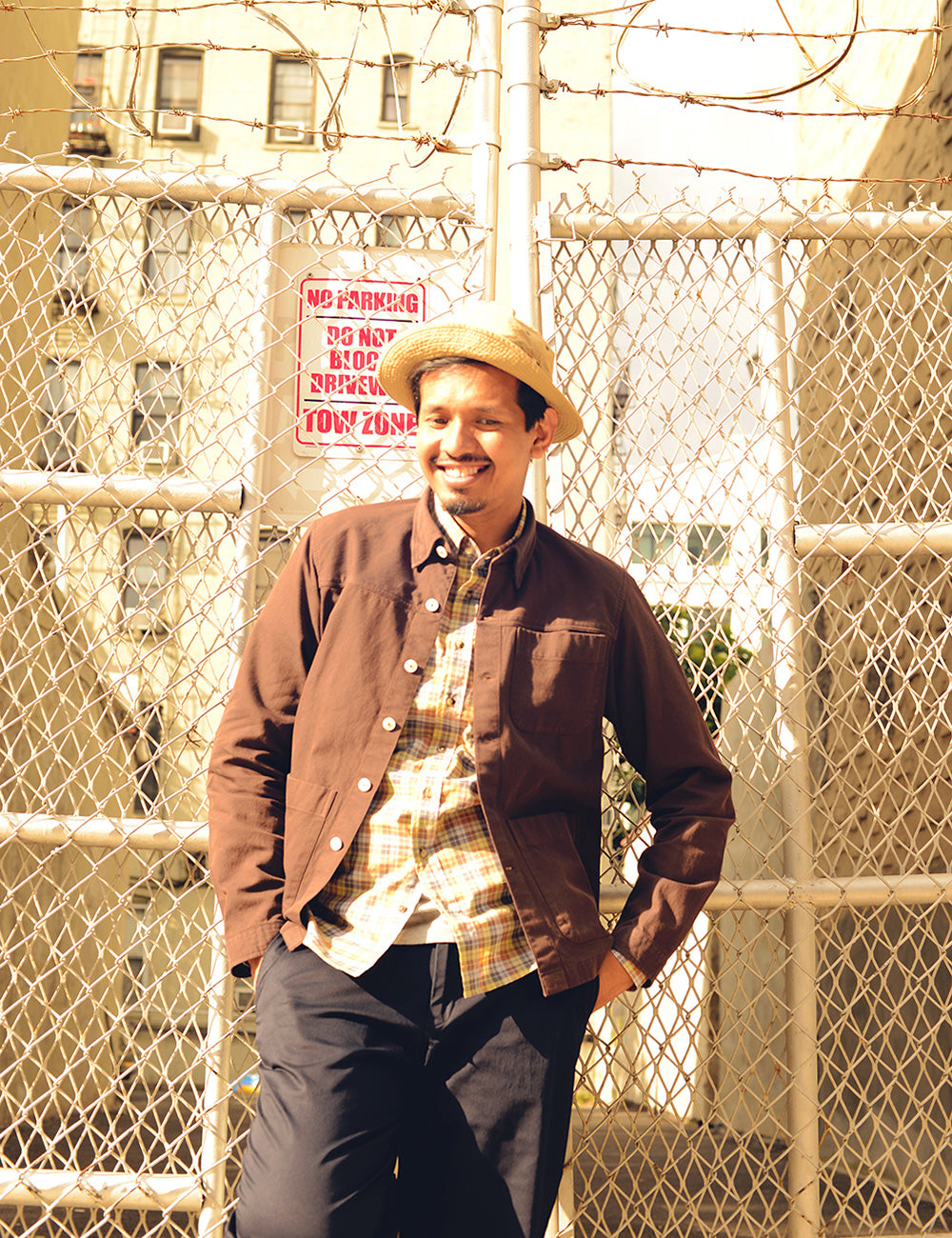 Model wears navy chinos, a plaid button-down shirt, brown cotton shirt jacket, and seersucker bucket hat. He is standing in front of a chain link fence.
