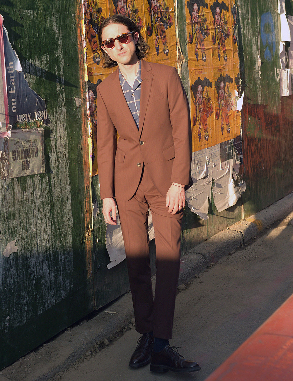 Model wears a rust/brown suit, blue check camp shirt, and sunglasses. He stands on a city street in front of a wall with posters and graffiti.