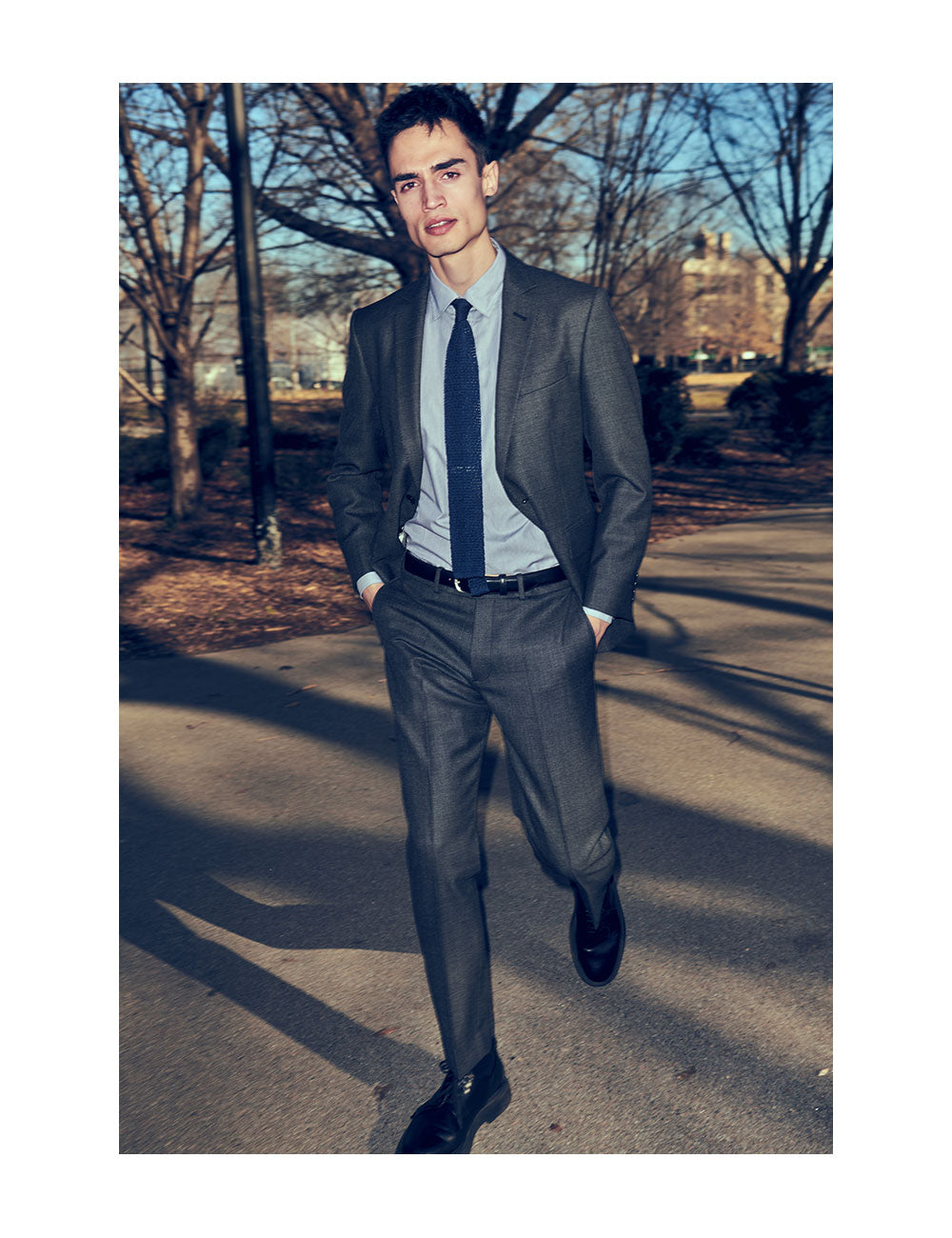 Model wearing charcoal suit, light blue dress shirt, and blue knit tie walking through a NYC park.