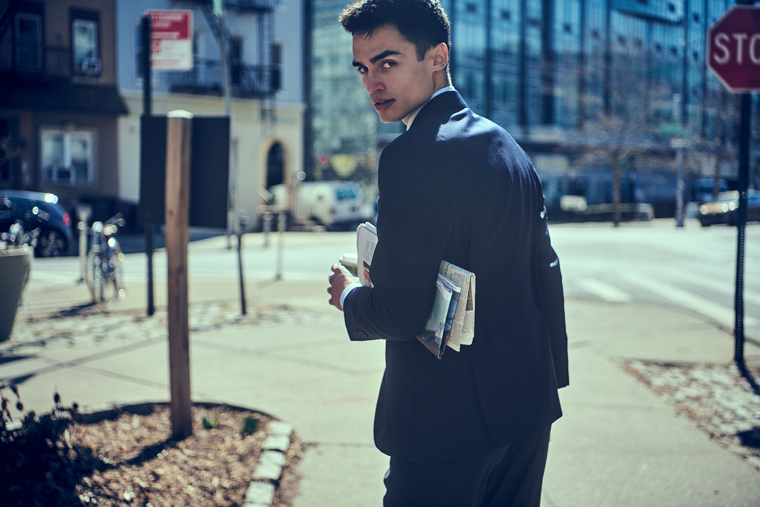 Model wears a navy suit. He is carrying coffee and a newspaper under his arm. He is shot from the back, looking over his shoulder.
