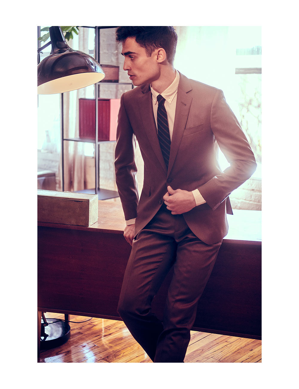 Model wears a brown suit, striped button down shirt, and striped tie. He is leaning against a wooden desk.