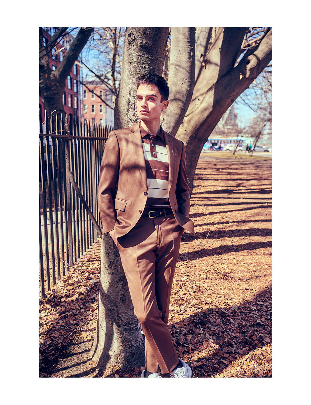Model is wearing a brown suit, brown belt, and brown and white striped polo shirt. He is standing in front of a tree in a NYC park.