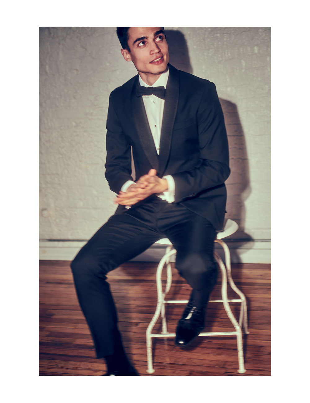 Model wearing a black tuxedo, white tuxedo shirt with dress studs, and black bow tie. He is sitting on a white stool.