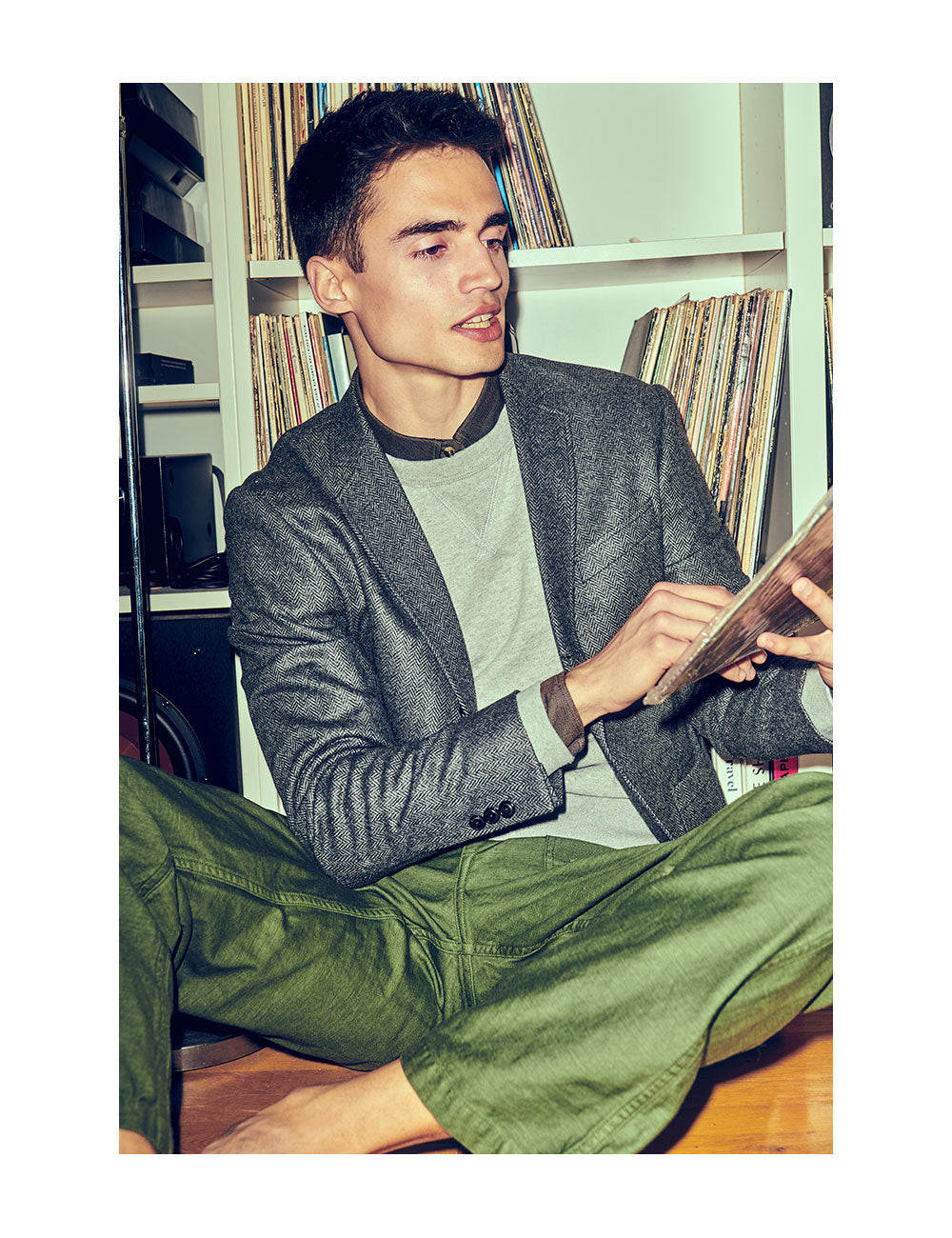 Model wearing a black button-down shirt, gray sweatshirt, charcoal herringbone unstructured blazer, and green chinos. He is sitting cross legged on the floor looking at records.