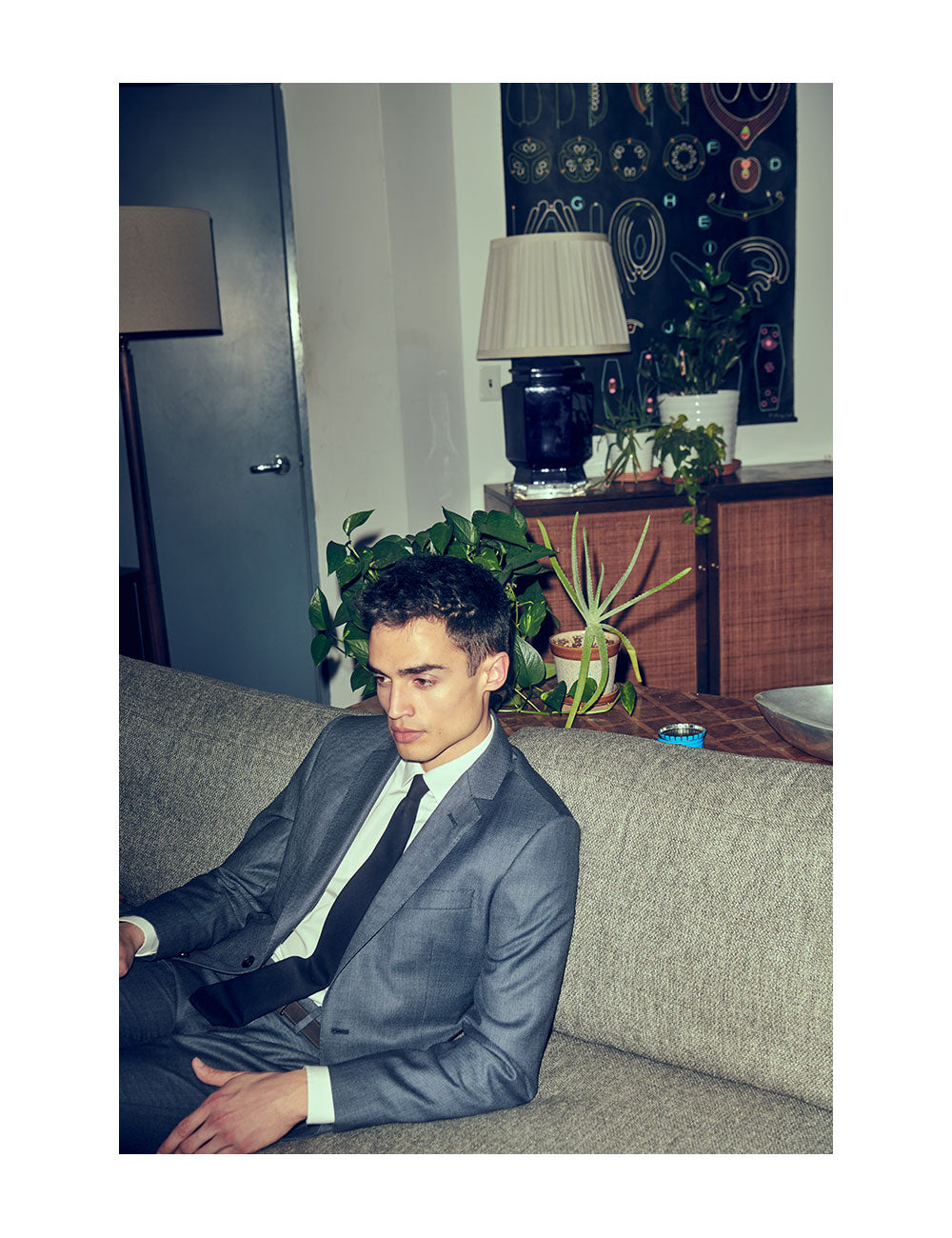 Model wearing charcoal suit, white dress shirt, dark blue knit tie, sitting on a couch in a NYC apartment.