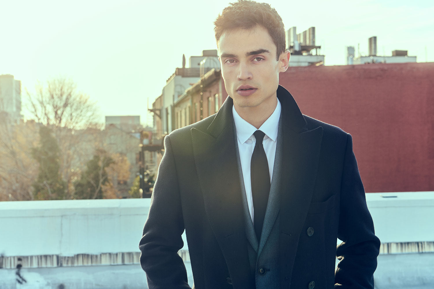 Model wears a charcoal suit jacket, white dress shirt, black tie, and black wool overcoat. He is standing on a NYC rooftop.
