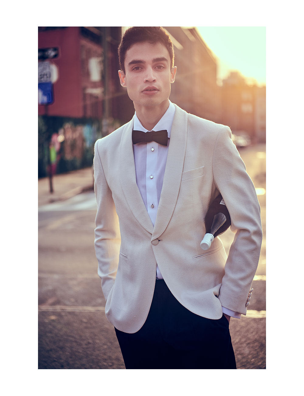 Model wearing an ivory dinner jacket, white dress shirt with mother of pearl shirt studs, black bow tie, and black tuxedo pants. He is holding a bottle of wine under his arm and standing on NYC street.