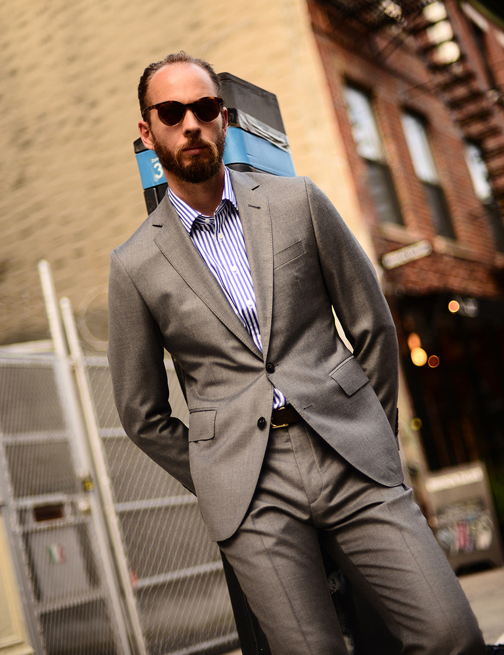 Model wears a gray suit jacket, white and navy striped dress shirt, and black belt. He is standing in front of a parking meter on a city street.