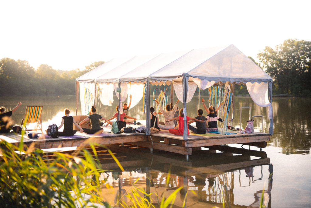 A group of people is participating in a yoga class on a floating platform on a serene lake at sunset. The platform is covered with a white tent that is open on the sides, adorned with colorful ribbons, creating a festive an