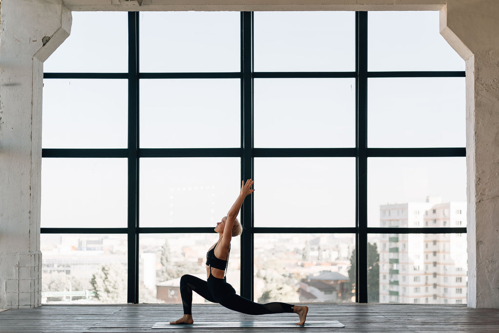 A person is performing a yoga pose in a spacious room with large windows that offer a view of the city skyline. The individual is in a lunge position with one knee on the yoga mat, the other leg extended back, and their arm