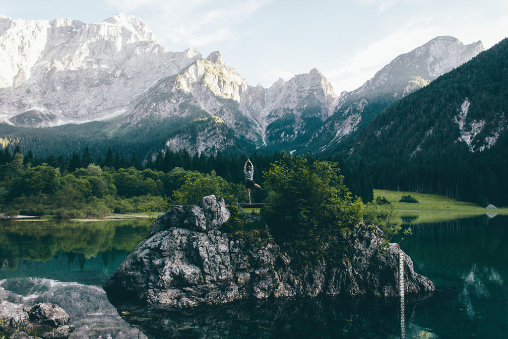 A serene scene where an individual is practicing yoga atop a large rock by a tranquil, reflective lake. The person is in the 'tree pose', balancing on one leg with the other foot against the standing leg's inner thigh, and