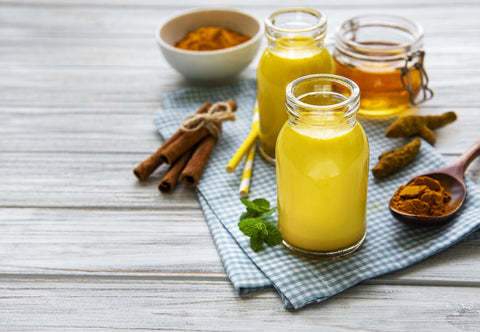 Two glass bottles of hemp infused golden milk on a placemat next to cinnamon sticks, a jar of honey, and a wooden spoon with powdered turmeric in it