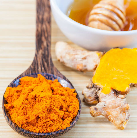 A wooden spoon with powdered turmeric alongside a piece of turmeric and a bowl of honey