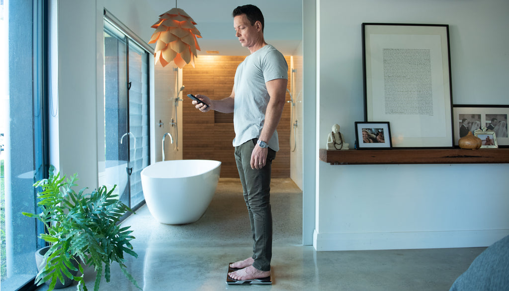 Man checking his measurements from his Tanita Body Composition Monitor on his phone in a bedroom on concrete floor.