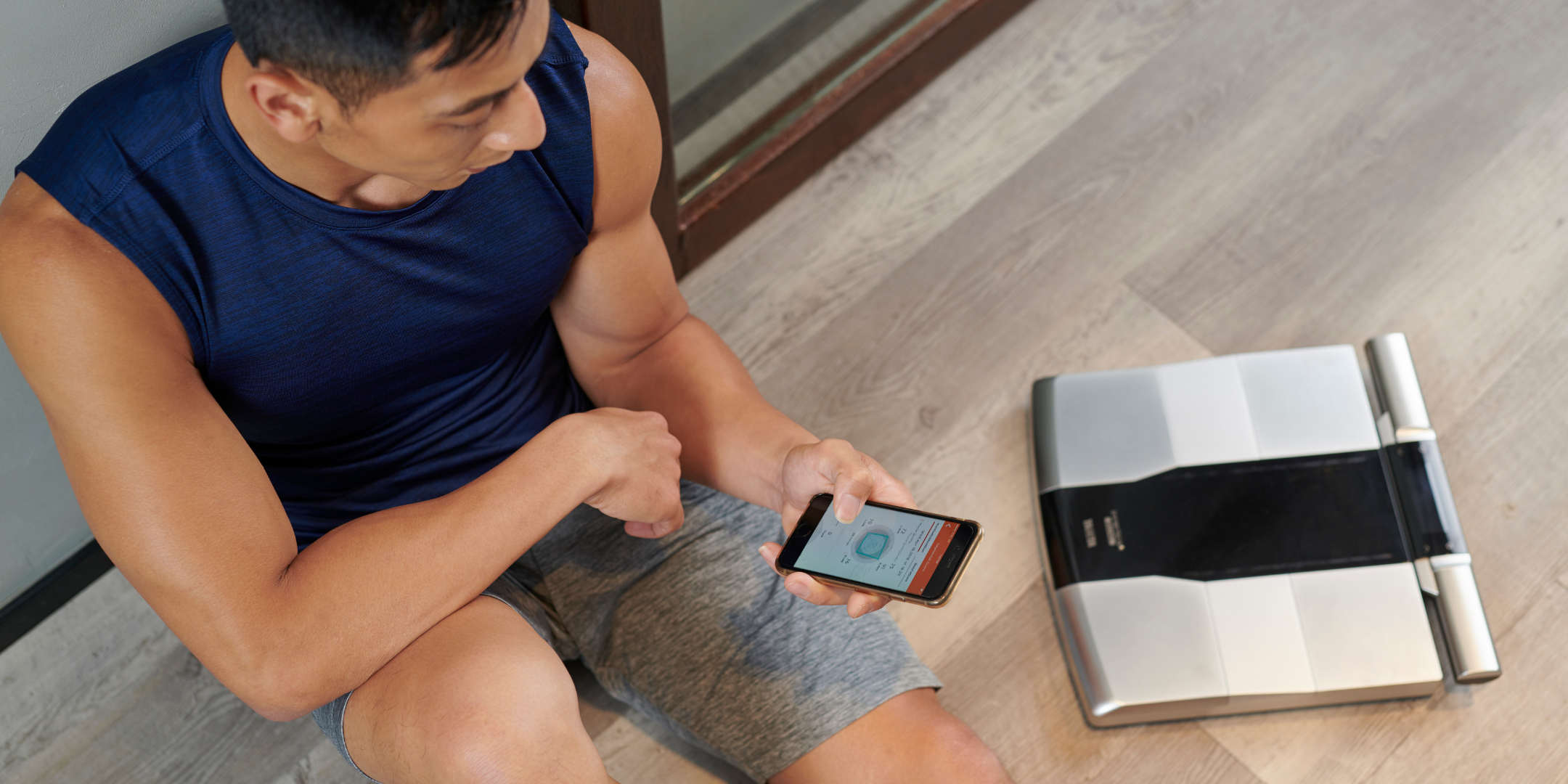Man sitting on the floor in a blue tank top reading body measurements in the Health Planet app next to his Tanita RD-545 Segmental Body Composition Monitor
