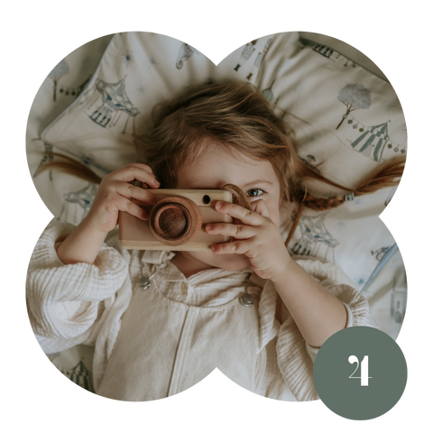 Toddler girl taking a photo with a wooden camera, laying on a circus tent printed pillow