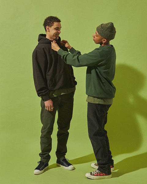 Two models stand in front of a green background. The model on the left wears the #5005 Workwear Pants in Vine Green, the #7006 T-shirt in Army Green and the #7004 Hooded Sweatshirt in Faded Black. The model on the right adjusts his sweatshirt. They wear the #7005 Sweatshirt in Vine Green, #7006 T-shirt in Army Green and dark denim jeans.