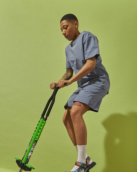 A model jumps on a pogo stick in front of a lime green background. They wear the #7007 Shorts in Teal and the #7006 T-shirt in Teal.