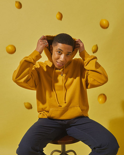 A model sits in front of a yellow background, wearing the #7004 Hooded Sweatshirt in Yellow and the #5005 Workwear Pants in Navy. He pulls his hood over his head. Around him are lemons, which appear as if they have been thrown.