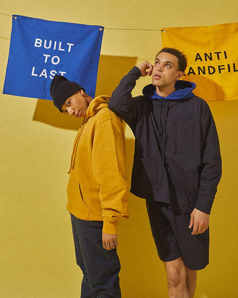 Two models stand in front of a yellow background. Two rectangular flags read “anti landfill” and “built to last”. The model on the left wears the #7004 Hooded Sweatshirt in Yellow and the #5005 Workwear Pants in Navy, along with a navy blue hat. The model on the right wears the #7004 Hooded Sweatshirt in Ultra Blue, the #3032 Tie Neck Smock in Midnight Blue and the #7007 Shorts in Navy.