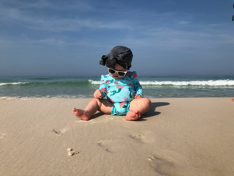 baby in cute swim suit and bathing hat sitting on the sand at the beach with sea behind