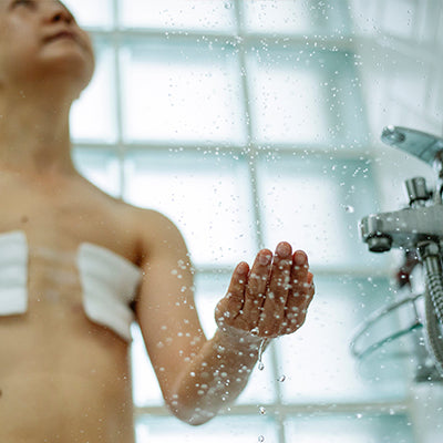 Woman holds her hand under shower stream, with dressings over mastectomy wounds