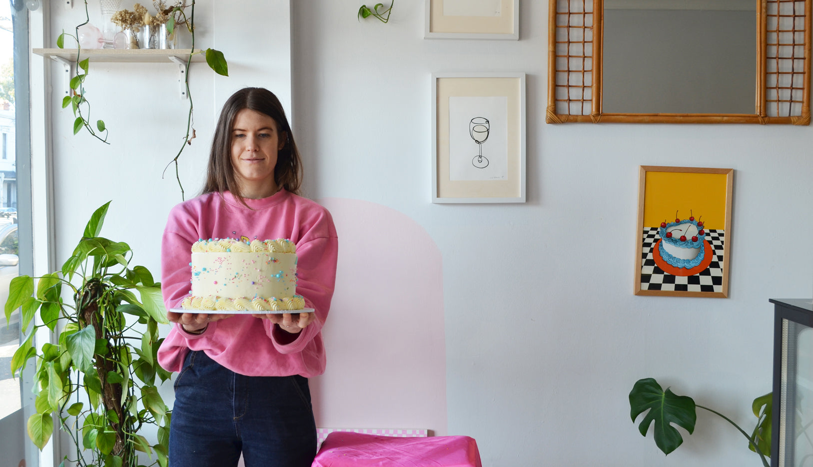 Matilda Chambers wearing a pink jumper holding a cream cake covered in sprinkles. A green plant sits in the background with prints on the wall behind her.