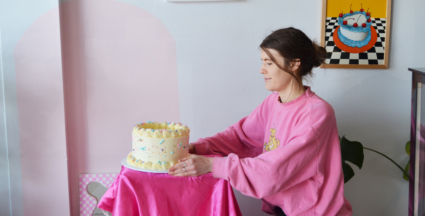Matilda Chambers sits in front of a cream and sprinkle cake wearing a pink jumper.